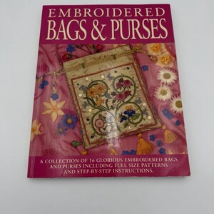 May include: A book titled "EMBROIDERED BAGS & PURSES" with a pink cover. The cover features an embroidered bag with floral designs and the text: "A COLLECTION OF 16 GLORIOUS EMBROIDERED BAGS AND PURSES INCLUDING FULL SIZE PATTERNS AND STEP-BY-STEP INSTRUCTIONS."