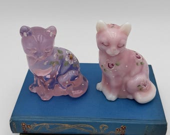 May include: Two ceramic cat figurines. One is a translucent purple with painted flowers, the other is pink with white accents and floral details. Both are seated with closed eyes. They rest on a stack of blue and green books.