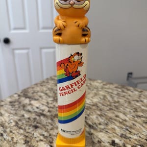 May include: Orange Garfield pencil case with a smiling Garfield figure on top. The cylindrical case is white with a rainbow stripe and the words "Garfield Pencil Case". A vintage collectible.