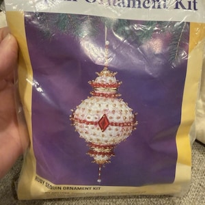 May include: A package labeled "Sequin Ornament Kit" featuring a detailed, ruby-colored sequin ornament. The ornament has a white base with red accents and a central ruby-colored diamond shape. The kit includes the ornament shape.
