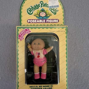 May include: A Cabbage Patch Kids poseable figure in its original packaging. The doll, named Jennifer Lillian, has a bald head, a white top with balloon graphics, and pink shorts and shoes. The box is yellow and green with First Edition text.