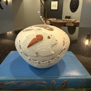 May include: A white ceramic decorative bowl with a textured frog design and red accents. The bowl sits atop a stack of blue and green books. The frog has a carrot-shaped nose. The bowl has cut-out designs.
