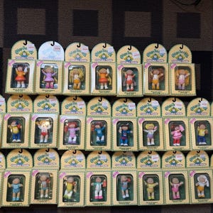 Peut inclure: Collection de figurines portables Cabbage Patch Kids dans leur emballage d'origine. Les figurines portent diverses tenues, avec des cheveux blonds, bruns et roux. L'emballage est beige avec des accents verts et jaunes.