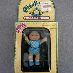 May include: A Cabbage Patch Kids poseable figure named Marna Jane in original packaging. The doll has a bald head, green eyes, and wears a light blue romper with a white diamond pattern and blue shoes. The packaging includes the text First Edition.