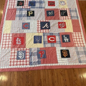 Könnte beinhalten: Eine Patchwork-Quilt mit verschiedenen Logos von Major League Baseball-Teams. Der Quilt hat einen roten Rand und besteht aus Quadraten in Rot, Weiß, Blau und Gelb. Zu den Logos gehören die Cardinals, Mets und Dodgers.