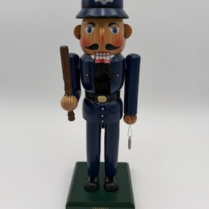May include: A wooden police officer nutcracker in a dark blue uniform and helmet, holding a baton. The nutcracker has a black mustache and beard, with a gold belt buckle. The base is green with the number "2006" on it.
