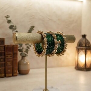 Puede incluir: Un conjunto de brazaletes de color verde oscuro con una superficie texturizada y pequeños adornos reflectantes. Los brazaletes están decorados con bordes de cuentas doradas y se exhiben en un soporte de barra horizontal de color dorado.