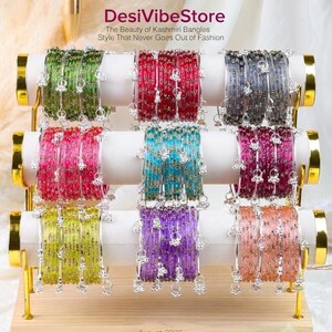 Może przedstawiać: Trzypoziomowy drewniany stojak ekspozycyjny z dziewięcioma zestawami kolorowych bransoletek w kolorach zielonym, czerwonym, szarym, różowym, turkusowym, magenta, limonkowym, fioletowym i brzoskwiniowym. Każda bransoletka ma srebrne zawieszki i napis DesiVibeStore August 2026.