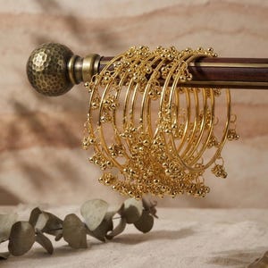 May include: A set of gold-toned metal bangles featuring small, dangling spherical charms attached to the thin circular bands. The bangles are displayed hanging on a horizontal wooden rod with a textured, dark metallic end cap.