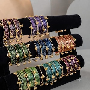 Pode incluir: Uma coleção de pulseiras brilhantes multicoloridas dispostas num suporte de exibição de veludo preto. Cada pulseira apresenta uma estrutura fina de metal dourado com pequenos pingentes dourados pendurados. As pulseiras estão agrupadas por cor.