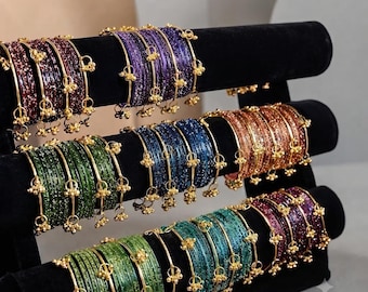 May include: A collection of multi-colored glittery bangles arranged on a black velvet display stand. Each bangle features a thin gold-tone metal frame with small dangling gold-tone charms. The bangles are grouped by color, including purple, green, blue, and copper.