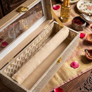 May include: A rectangular, champagne-coloured jewellery box with a hinged lid featuring a clear window with the name Georgia etched in white script. The interior is lined with a soft, beige velvet fabric and includes a single cylindrical roll.