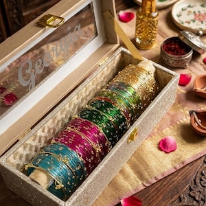 Puede incluir: Un joyero rectangular dorado con purpurina y tapa transparente que muestra el nombre Georgia en letra cursiva. En su interior, una colección de brazaletes apilados en tonos verde azulado, magenta, verde y dorado con detalles en tono dorado.
