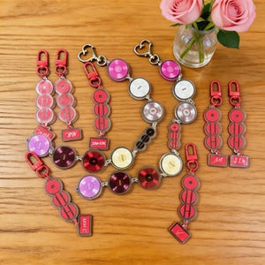 May include: A collection of keychains featuring red clasps and circular charms in various colours, designed to look like miniature vinyl records. Some keychains have name tags. A vase of pink roses is in the background.