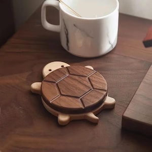 Könnte beinhalten: Ein hölzerner Untersetzer in Schildkrötenform mit einem dunkelbraunen, geometrischen Schalendesign. Der Untersetzer liegt auf einer dunklen Holzoberfläche neben einer weißen Tasse mit Marmormuster.