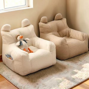 Puede incluir: Dos sillones puff de felpa en crema y beige. Cada silla tiene un respaldo redondeado con dos protuberancias en forma de oreja en la parte superior. La silla crema tiene un pequeño bolsillo lateral y un ganso de peluche. Las sillas están sobre una alfombra clara.