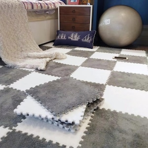 Puede incluir: Baldosas entrelazadas de espuma para el suelo en un patrón de tablero de ajedrez blanco y gris. Los cuadrados grises tienen una textura suave y afelpada. Varias baldosas están apiladas, mostrando los bordes entrelazados. Se ve un cojín azul marino con un diseño de barco.