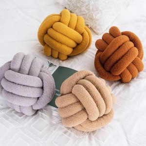 May include: Four decorative knot pillows in shades of yellow, brown, lavender, and beige. The pillows are made of a soft, textured material and are arranged on a white surface. The knot design adds a unique touch to the home decor.