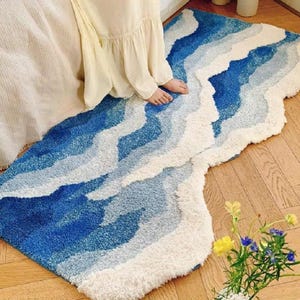 May include: A blue and white rug with a wavy design, resembling ocean waves. The rug is made of a soft, textured material and is placed on a wooden floor. The rug's color palette includes shades of blue and white.