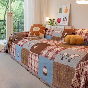 Puede incluir: Una funda de sofá patchwork multicolor con una mezcla de cuadros, lisos y cuadrados estampados. La funda presenta imágenes bordadas de perros en monopatines, cupcakes y estrellas. Un cojín decorativo de flores y un cojín con forma de calabaza también están en el sofá.