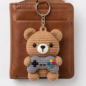 May include: A crocheted brown bear keychain with a gray video game controller. The bear has black eyes, a white muzzle, and pink cheeks. The controller has colorful buttons. The keychain is attached to a brown leather wallet.