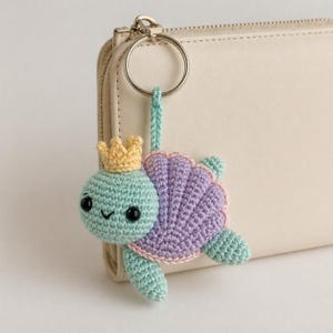 Puede incluir: Un llavero de tortuga hecho a mano con ganchillo. La tortuga tiene una cabeza azul claro con una corona amarilla, un caparazón morado y aletas azul claro. El llavero está sujeto a un anillo plateado y una correa azul claro.