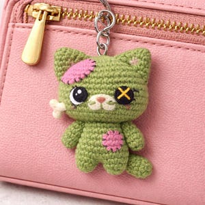 May include: A green crocheted cat keychain with pink patches and a bone. The cat has one black eye, an X for an eye, and a pink patch on its chest. The keychain is attached to a silver chain and a pink zippered wallet.