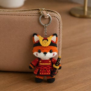 Könnte beinhalten: Gehäkelter Fuchsschlüsselanhänger in Orange, Rot, Schwarz und Weiß, gekleidet als Samurai. Der Fuchs hat ein kleines Schwert und ist an einem silbernen Schlüsselring befestigt. Der Schlüsselanhänger befindet sich neben einer beigen Geldbörse.