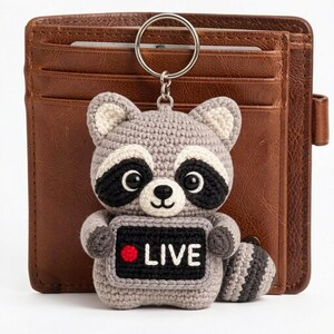 May include: Crocheted raccoon keyring with a silver ring. The raccoon is grey, black, and white, holding a sign that says "LIVE" with a red dot. The keyring is in front of a brown leather wallet.