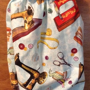 May include: A light blue drawstring bag with a repeating pattern of sewing machines, measuring tapes, scissors, and buttons. The bag has a ruffled top and a brown ribbon drawstring.