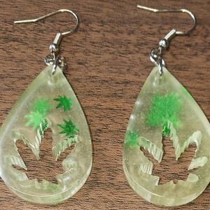 Könnte beinhalten: Ein Paar transparente Harzohrringe mit einem grünen Marihuana-Blatt-Design. Die Ohrringe sind tropfenförmig und haben silberne Haken.