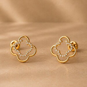 May include: A pair of gold stud earrings shaped like four-leaf clovers. Each earring features a polished gold outline with a smaller inner clover shape encrusted with small, clear, sparkling stones. The earrings have secure screw-back posts.