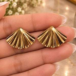 May include: A pair of gold-toned stud earrings shaped like fan-like seashells. Each earring features a polished, reflective surface with distinct vertical ridges radiating from the center point, creating a textured, pleated appearance.