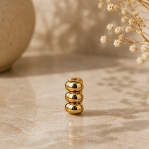 May include: A gold-toned ear cuff featuring three stacked, smooth, rounded, bead-like segments. The metal has a polished, reflective finish that catches the light, creating a bright, metallic appearance with distinct highlights on each rounded section.