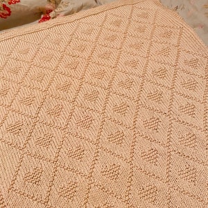 May include: A pale pink knitted blanket with a diamond pattern. The blanket is made with a soft yarn and has a subtle texture.