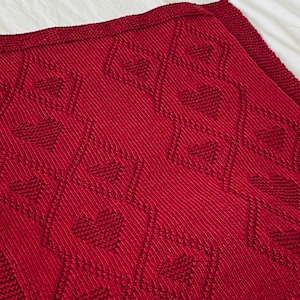 Può includere: Una coperta di maglia rossa con un motivo a cuore ripetuto. La coperta è realizzata con un filato morbido e testurizzato.