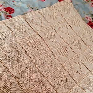 May include: A cream-colored knitted blanket featuring a repeating grid pattern of textured hearts. Each heart is formed by a distinct knit stitch pattern within a square border, creating a uniform and structured design across the entire fabric surface.