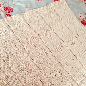 May include: A cream-colored knitted blanket featuring a repeating grid pattern of textured hearts. The blanket is constructed with a garter stitch border and vertical and horizontal lines separating each individual heart square design.