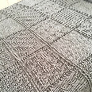 May include: A grey knitted blanket with a textured, geometric pattern. The blanket features a variety of stitched squares and rectangles, creating a patchwork design. The yarn appears soft and the blanket is likely for a bed or couch.