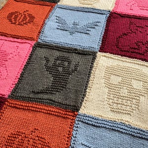 May include: A knitted Halloween-themed blanket featuring squares with various designs. The blanket showcases a pumpkin, bat, ghost, and skull in orange, blue, gray, and cream colors. The blanket is a cozy, handmade item.