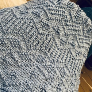 Op de afbeelding: Een close-up van een blauwe gebreide deken met een geometrisch patroon. De deken is gemaakt van zacht garen en heeft een subtiele glans.