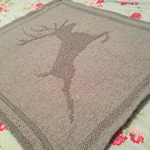 May include: A gray knitted blanket with a deer design. The blanket has a border around the edges.