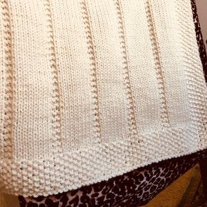 Può includere: Una coperta di maglia bianca con una trama a coste. La coperta è piegata e drappeggiata su una sedia.