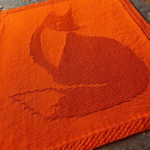 May include: A knitted orange blanket with a fox silhouette design. The blanket is made with a soft, textured yarn and has a simple, modern design.