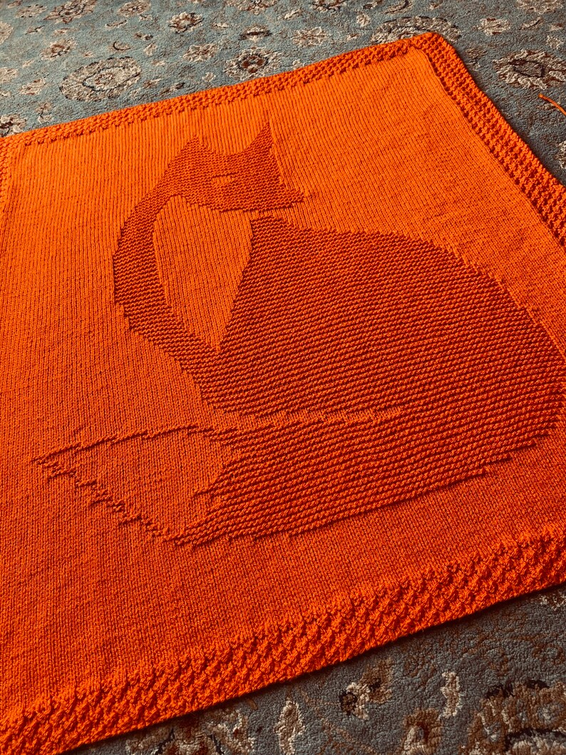 May include: A knitted orange blanket with a fox silhouette design. The blanket is made with a soft, textured yarn and has a simple, modern design.
