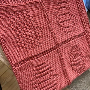 May include: A square, terracotta-colored knitted blanket featuring four distinct textured panels. Each panel displays a unique raised pattern, including a heart, a pumpkin, a bee, and a tree, all created with consistent knit and purl stitches.