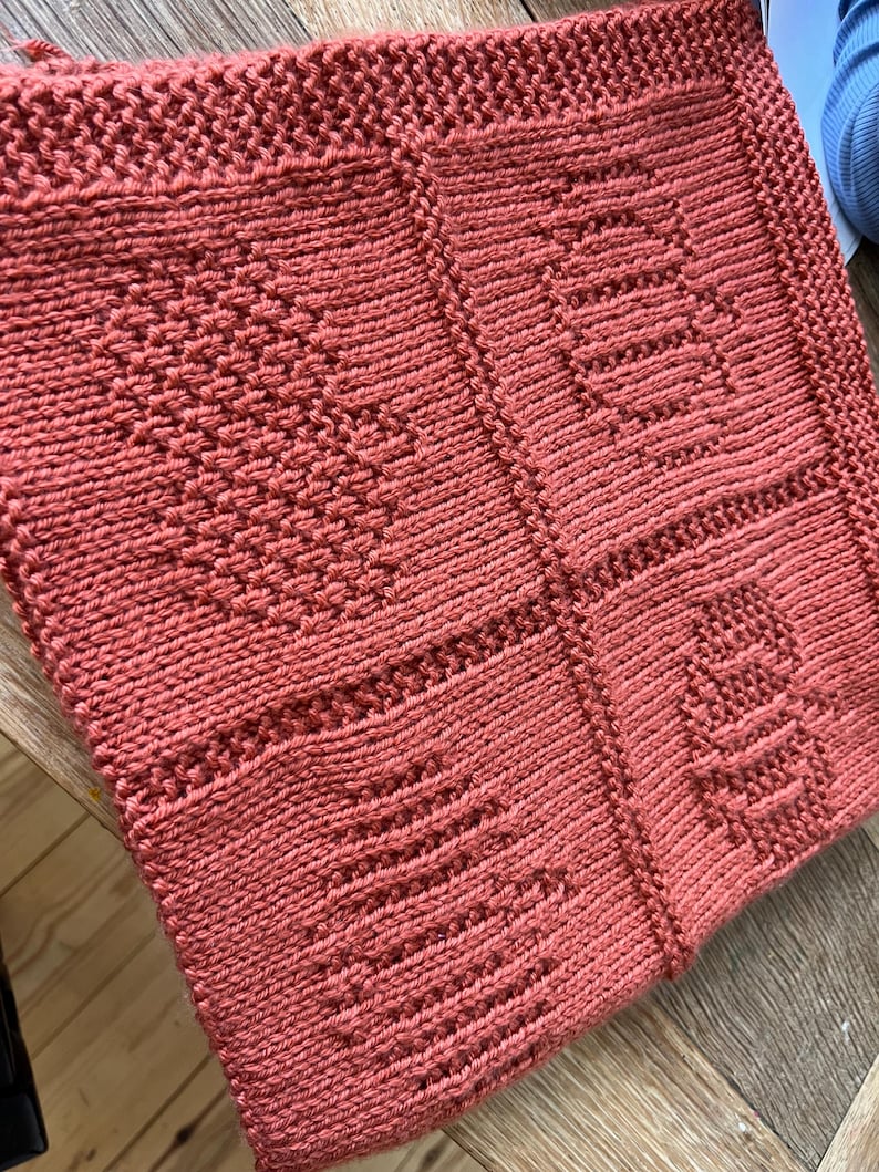 May include: A square, terracotta-colored knitted blanket featuring four distinct textured panels. Each panel displays a unique raised pattern, including a heart, a pumpkin, a bee, and a tree, all created with consistent knit and purl stitches.