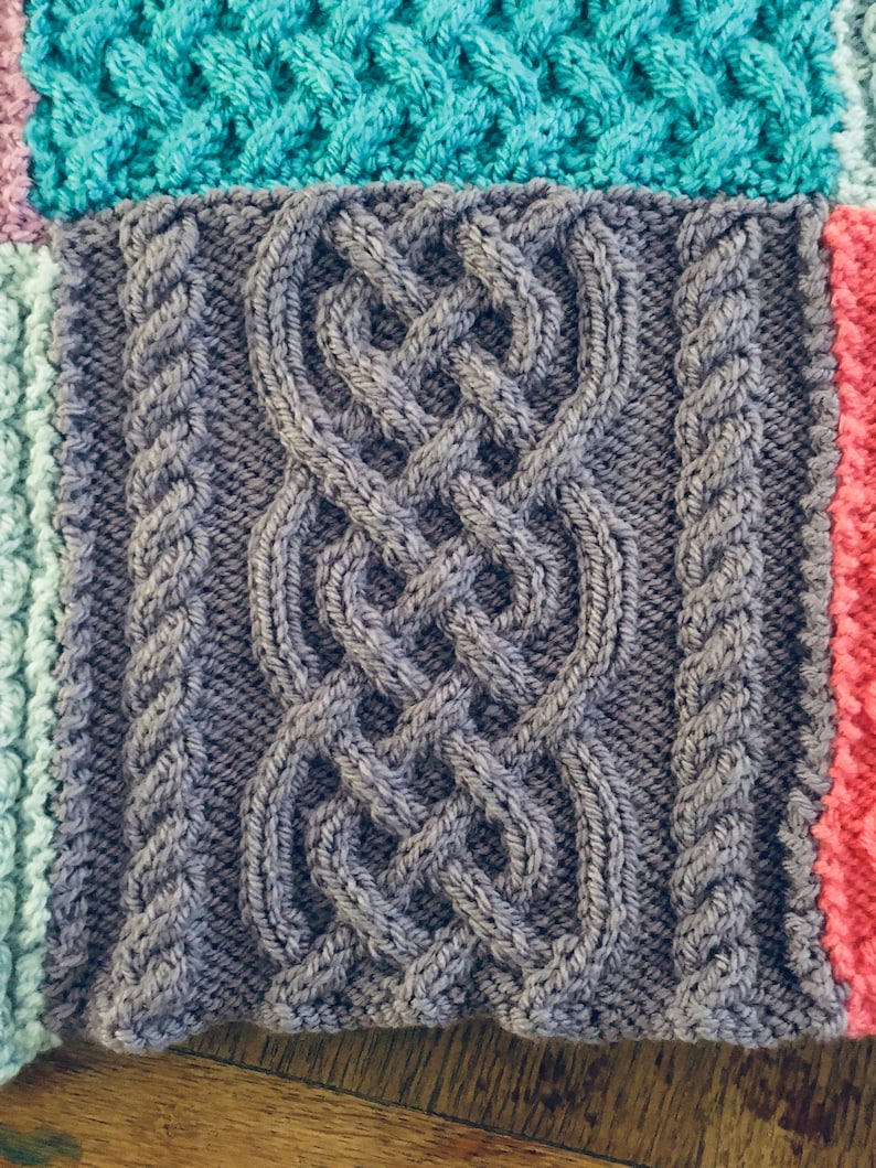 Peut inclure: Gros plan sur une couverture tricot&eacute;e avec un motif en c&acirc;ble gris. La couverture est compos&eacute;e de carr&eacute;s de diff&eacute;rentes couleurs, dont le bleu sarcelle, le rose et le bleu clair.