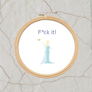May include: A cross-stitch embroidery hoop featuring the phrase "F*ck it!" in blue letters. The design includes a pixelated image of a woman in a blue dress with blonde hair. The hoop is made of light brown wood.