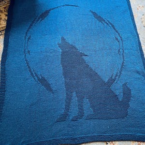 Könnte beinhalten: Eine gestrickte blaue Decke mit einer dunkelblauen Silhouette eines heulenden Wolfes vor einem Vollmond. Die Decke hat ein strukturiertes Aussehen und einen dunkelblauen Rand. Das Wolf- und Mond-Design ist deutlich auf der Decke zu sehen.